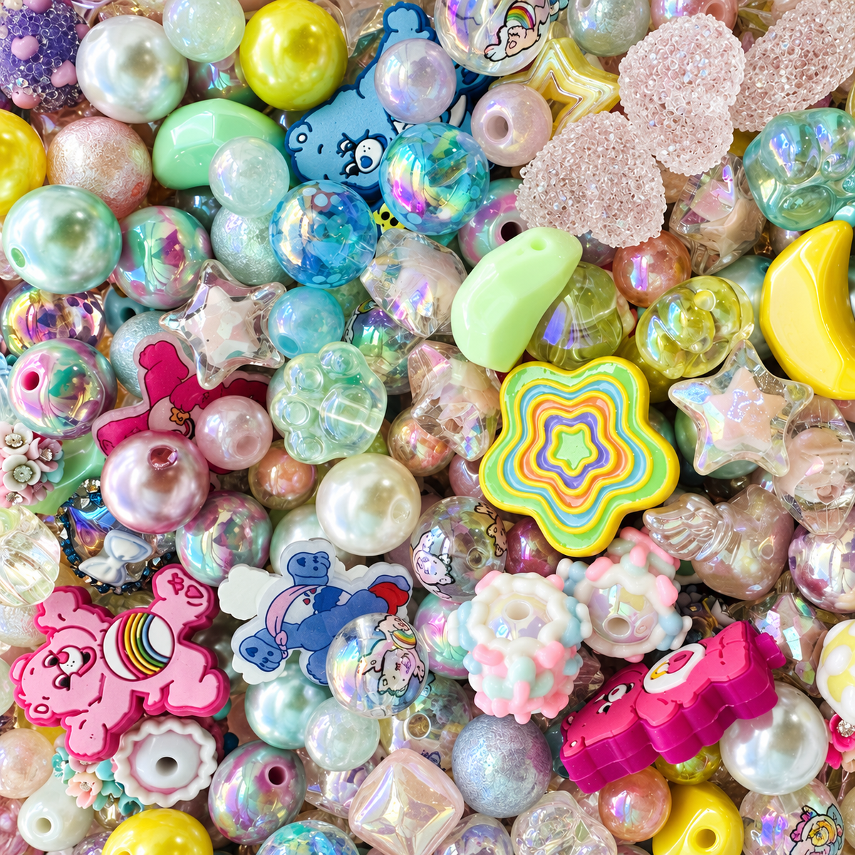 Close-up of Care Buddies bead mix showing pastel pink, aqua, mint, lavender and yellow beads with pearl, iridescent, transparent and textured finishes, plus stars, moons, rainbow details, floral accents and colorful character focal pieces.