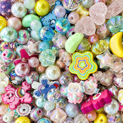 Close-up of Care Buddies bead mix showing pastel pink, aqua, mint, lavender and yellow beads with pearl, iridescent, transparent and textured finishes, plus stars, moons, rainbow details, floral accents and colorful character focal pieces.