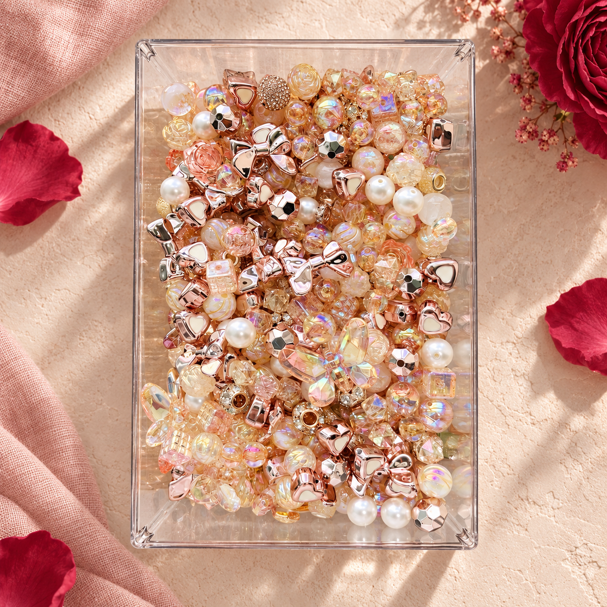 Overhead view of Rose Gold bead mix in a clear tray featuring metallic rose-gold bows, creamy pearl beads, champagne iridescent rounds, faceted crystal-look beads and warm blush accents surrounded by rose petals.