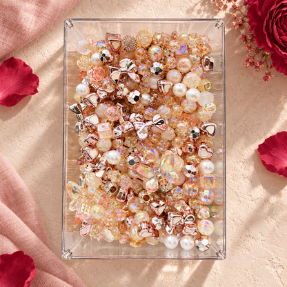 Overhead view of Rose Gold bead mix in a clear tray featuring metallic rose-gold bows, creamy pearl beads, champagne iridescent rounds, faceted crystal-look beads and warm blush accents surrounded by rose petals.