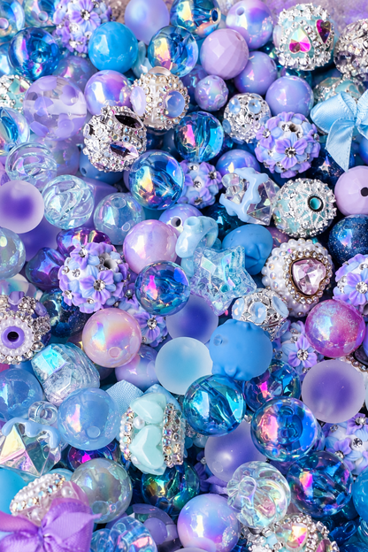 Overhead view of Periwinkle Dreams bead mix featuring periwinkle, lavender, aqua and blue beads with iridescent rounds, floral beads, bows, rhinestone accents and decorative focal beads.