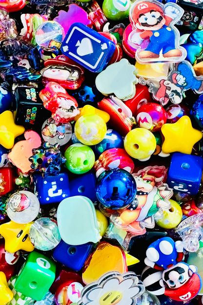 Close-up of the Retro Gamer bead mix showing blue question-mark cubes, green game-symbol blocks, character-inspired focal pieces, red hearts, glossy blue rounds, translucent beads, colorful game icons and dimensional arcade-inspired shapes.