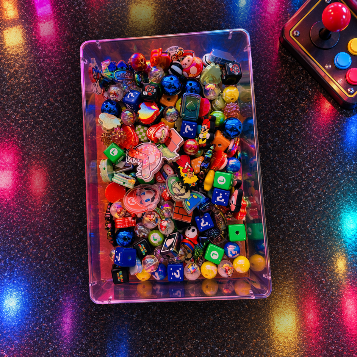 Overhead view of Retro Gamer beads in a clear tray on a glowing arcade-style surface, showing blue question-mark cubes, green game-symbol blocks, colorful character-inspired focal pieces, glossy rounds, stars and other gaming-themed shapes.