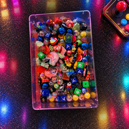 Overhead view of Retro Gamer beads in a clear tray on a glowing arcade-style surface, showing blue question-mark cubes, green game-symbol blocks, colorful character-inspired focal pieces, glossy rounds, stars and other gaming-themed shapes.