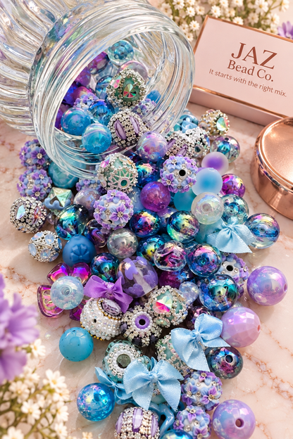 Periwinkle Dreams bead mix spilling from a clear jar, showcasing lavender and blue floral beads, iridescent rounds, sparkling rhinestone focal beads, stars and soft blue and purple bow accents.