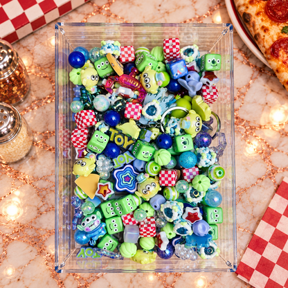 Close-up of Pizza Planet bead mix showing lime-green three-eyed alien-inspired focal beads, smiling green character cubes, red-and-white checkerboard beads, deep blue rounds, aqua and iridescent beads, a pizza slice, layered star, striped rounds and other playful dimensional shapes.
