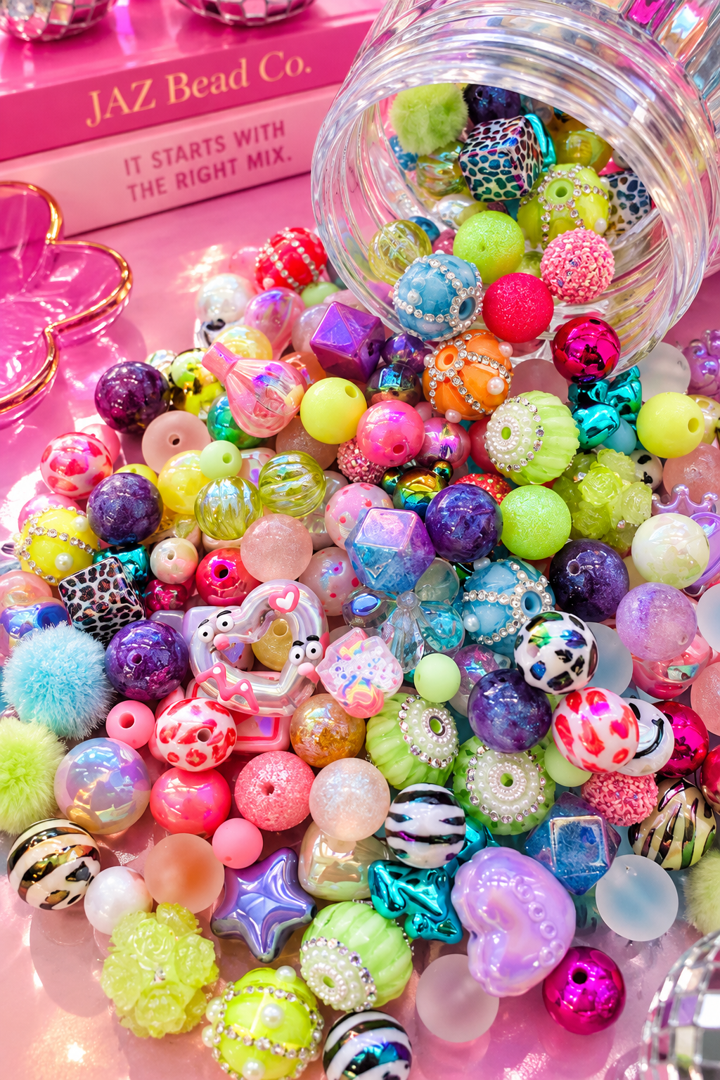 Clear jar of Cosmic Crush tipped on its side with colorful beads spilling across a glossy pink surface, including bright rounds, animal-print cubes, iridescent geometric beads, rhinestone accents and whimsical focal pieces in pink, purple, aqua, lime and orange.