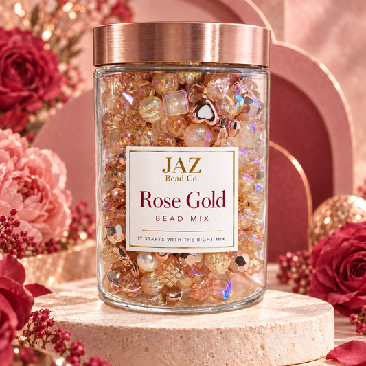JAZ Bead Co. Rose Gold bead mix in a clear jar with a rose-gold metallic lid, filled with rose-gold bows, champagne iridescent beads, creamy pearls and faceted accents, displayed among blush and deep rose flowers.