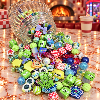 Overhead view of Pizza Planet bead mix in a clear tray featuring green three-eyed alien-inspired characters, smiling green cubes, blue and aqua round beads, red-and-white checkerboard cubes, stars, striped beads and colorful novelty focal pieces beside pizza-parlor accents.