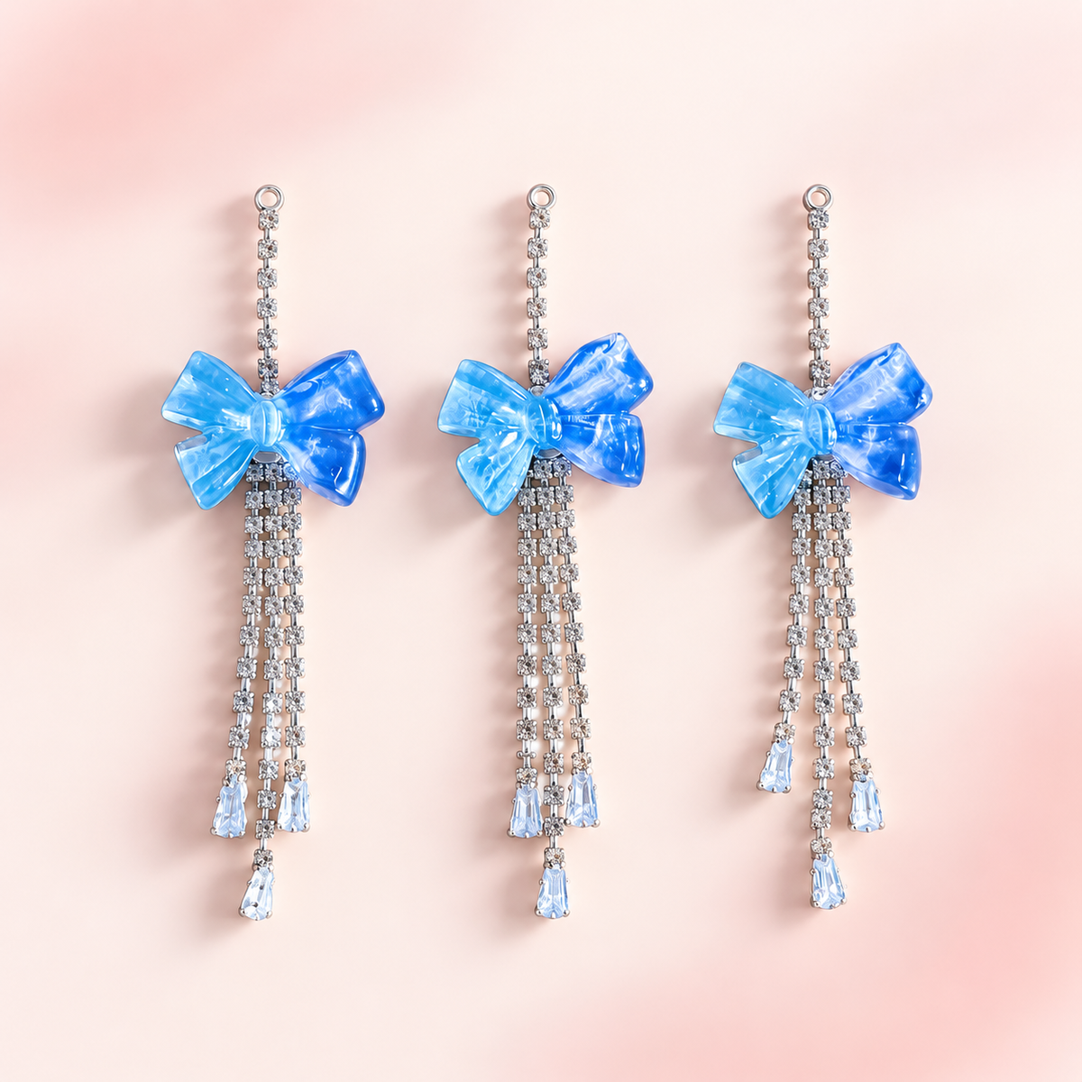 Three blue translucent bow dangle charms with silver rhinestone chains and light blue crystal teardrop accents displayed on a soft blush pink background.
