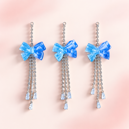 Three blue translucent bow dangle charms with silver rhinestone chains and light blue crystal teardrop accents displayed on a soft blush pink background.